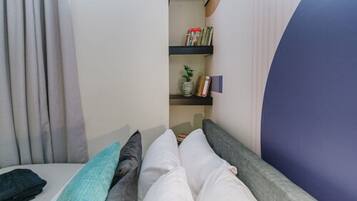 A bedroom with a bed, pillows, a nightstand, a shelf with books and a vase, and a window with curtains.
