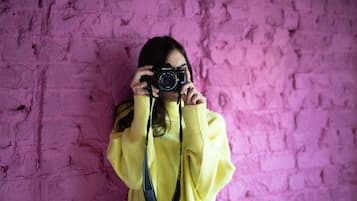 Eine Person hält eine Nikon Kamera mit Trageband vor einer rosafarbenen Ziegelwand.