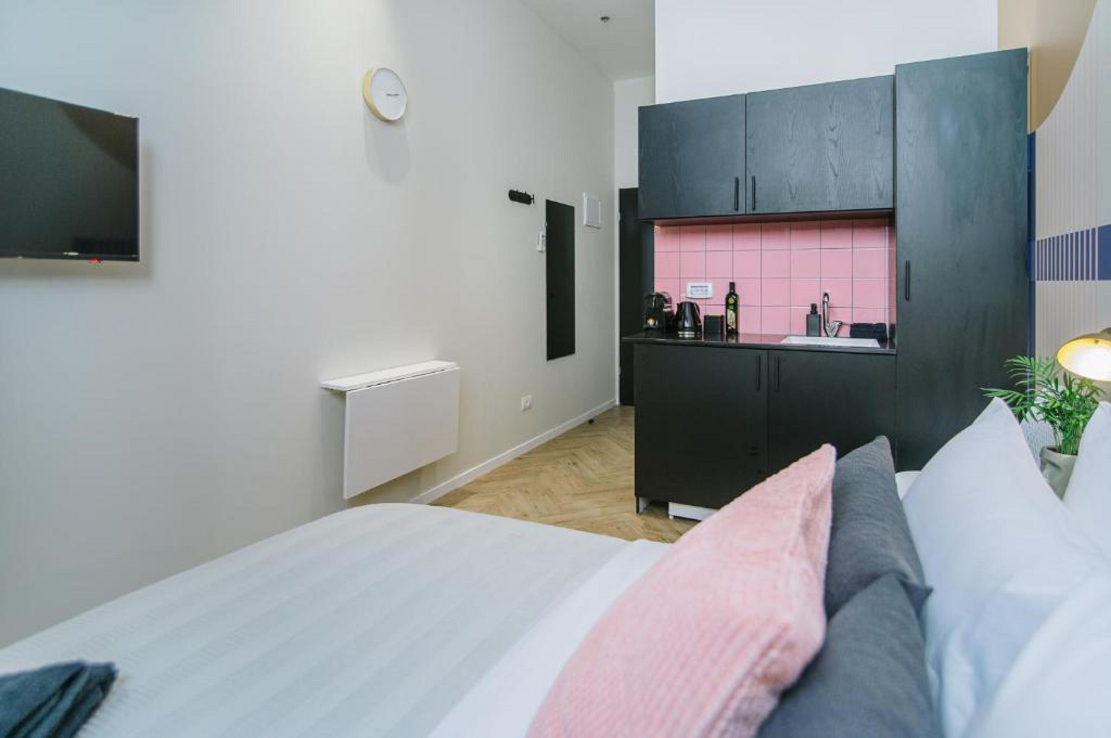A compact hotel room with a bed, a small kitchenette, a TV, and a radiator.