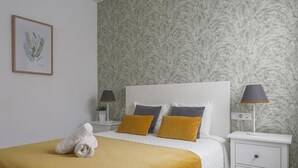 A neatly made bed with a white headboard, two pillows, and a mustard-colored throw blanket. There is a small bedside table with a lamp and a framed picture on the wall. - Pure Sevilla - Arenal - 2 bed common terrace 5 px (Sevilla)