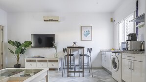 A modern kitchen with a built-in washing machine, a dining area with a table and chairs, a TV mounted on the wall, and a potted plant. - Pure Sevilla - Arenal - 2 bed common terrace 5 px (Sevilla)