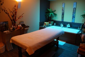 Een massagetafel in een kamer met een groot bad, een klein plantje en kunst aan de muur.