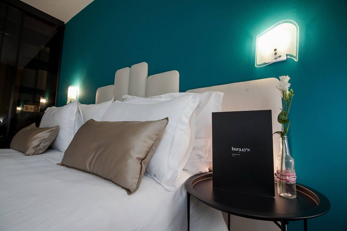 Un lit bien fait, avec des oreillers blancs et beiges, une table de chevet noire ornée d’un vase de fleurs, et une boîte noire portant l’inscription « bura45N » accrochée au mur.