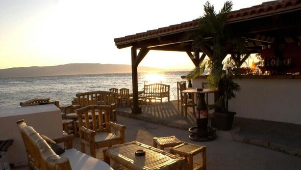 Une terrasse au bord de la plage, avec des meubles en bois, un palmier et une vue sur la mer au coucher du soleil.