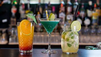 Tres bebidas distintas sobre una barra, con un fondo borroso de botellas y vasos.