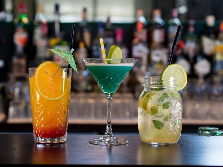Tres bebidas distintas sobre una barra, con un fondo borroso de botellas y vasos.