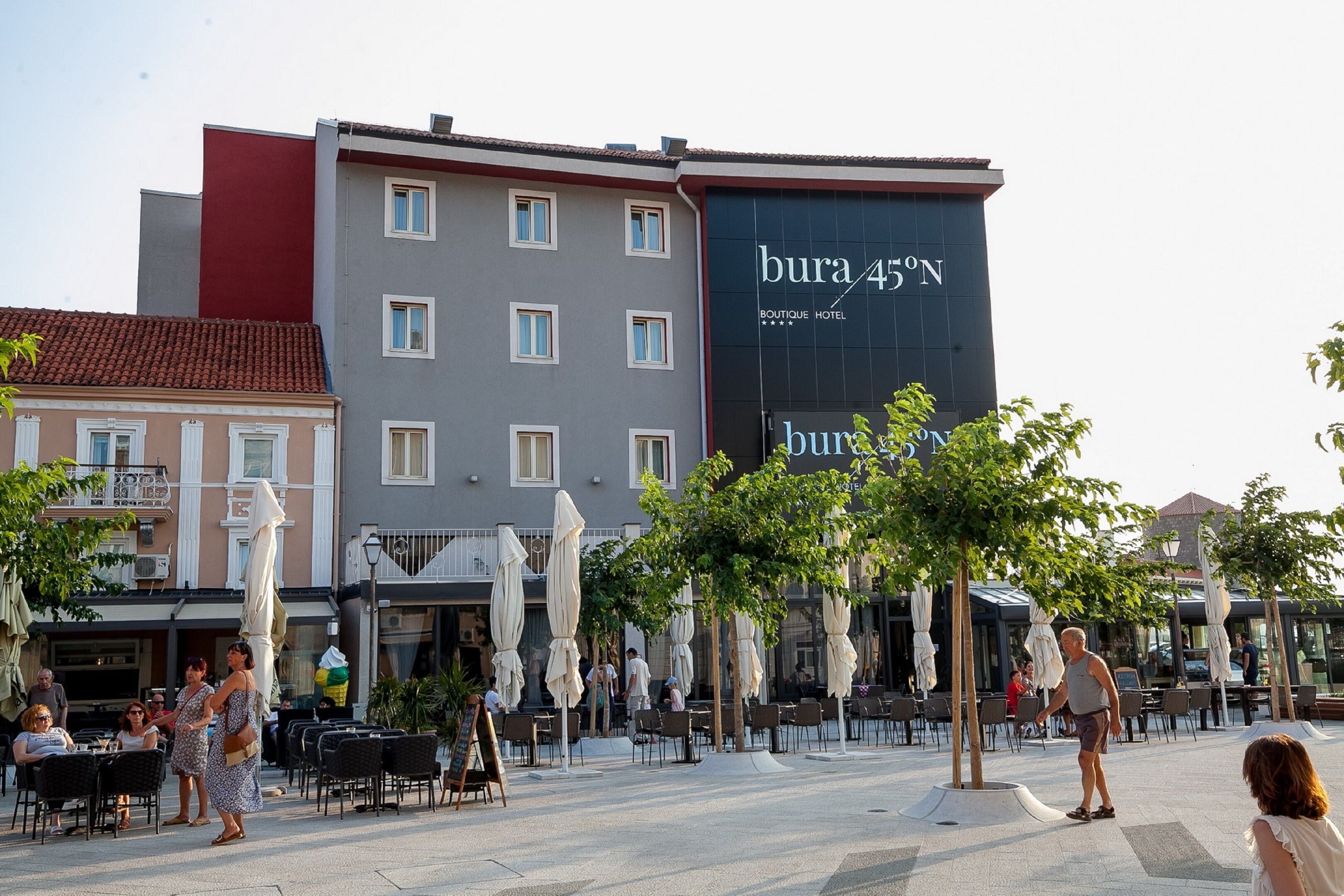 An outdoor cafe area with tables and chairs, a building with 'bura 45'N' signage, and a few people.