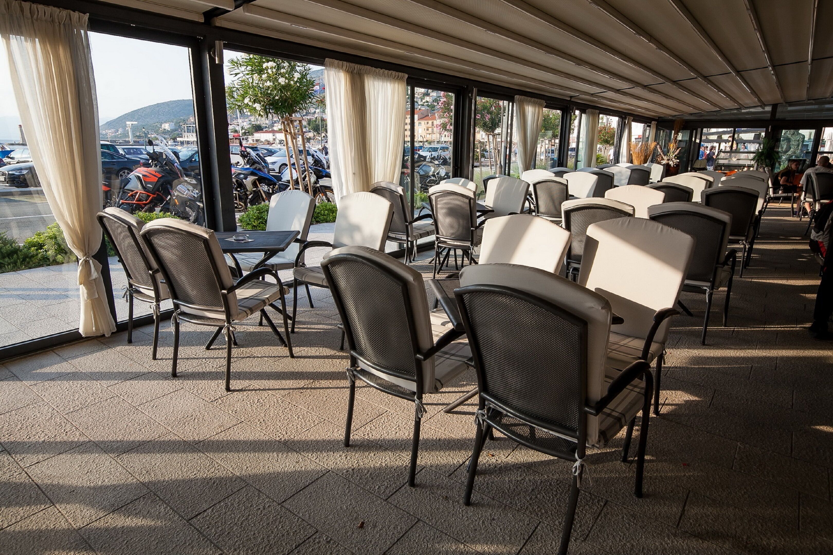 A covered outdoor seating area with tables and chairs, large windows with curtains, and a view of parked vehicles and buildings.