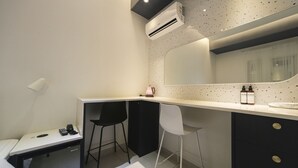 Una kitchenette compacta con un refrigerador pequeño, un lavamanos y un espejo sobre la encimera. Hay dos taburetes y una cama con una almohada blanca.