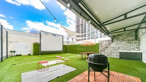 Un área de patio exterior con césped artificial, una terraza de madera, una parrilla y una carpa blanca.