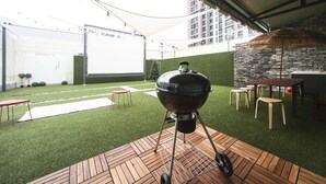 Una parrilla para barbacoa sobre un muelle de madera con césped artificial, un muro de piedra y una valla blanca al fondo.
