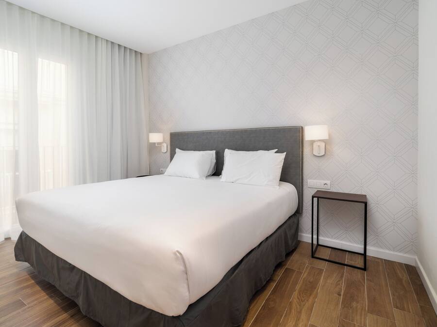 A neatly made bed with a grey headboard, white bedding, and a small bedside table with a lamp.