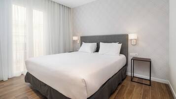 Cama bem arrumada com cabeceira cinza, roupa de cama branca e uma pequena mesa de cabeceira com abajur.