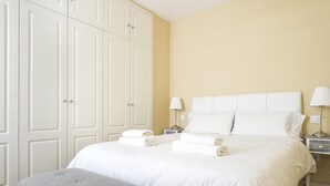 Une chambre d’hôtel avec un lit, un banc gris, des rideaux blancs et une armoire.