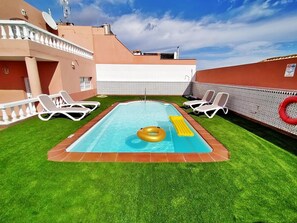 A rectangular swimming pool with a yellow inflatable ring, surrounded by artificial grass and white lounge chairs.