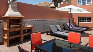 Een terras buiten met een bakstenen barbecue, lounge stoelen en een palmboom.