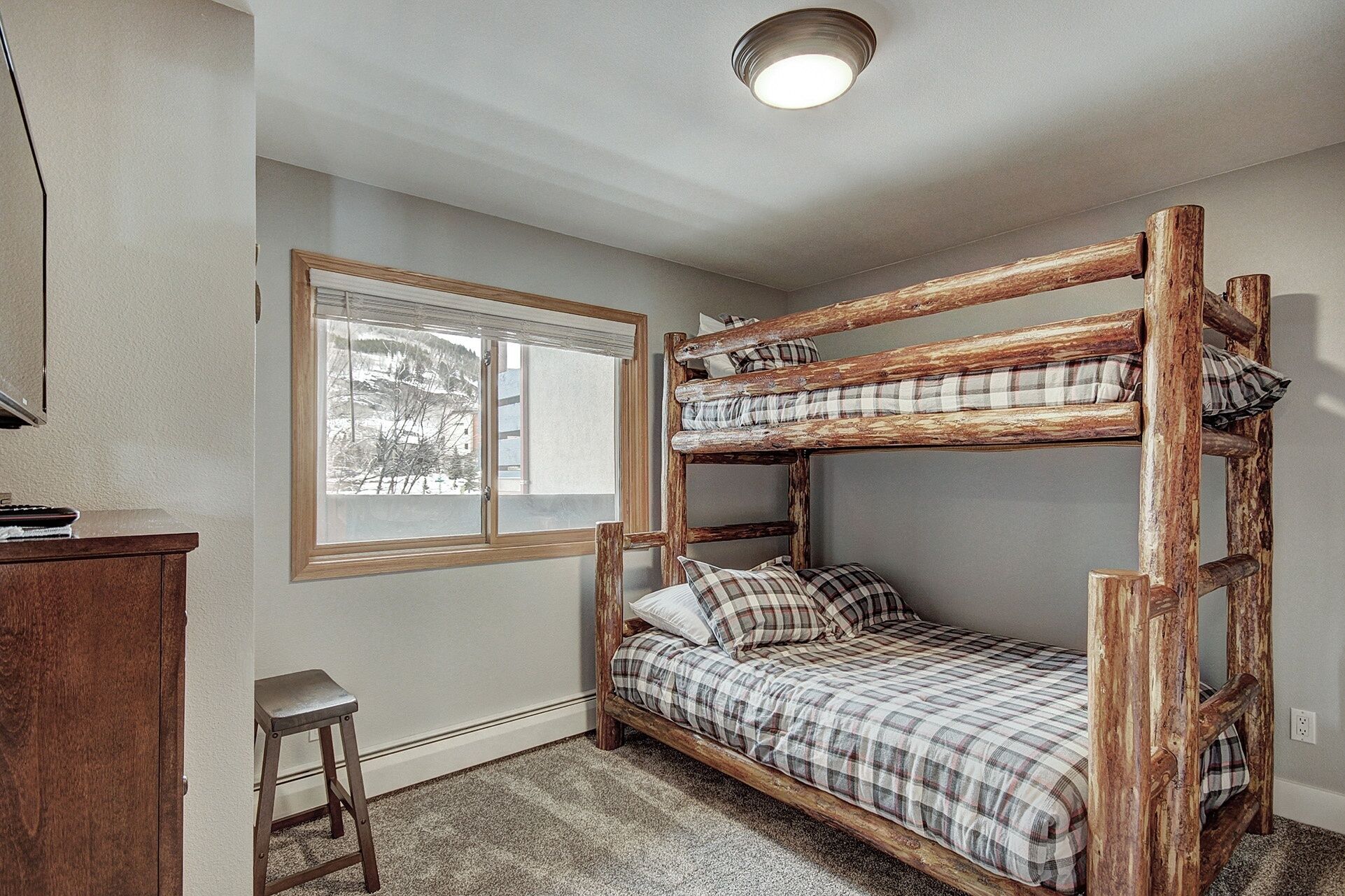 Una cama litera con ropa de cama escocesa, un taburete de madera, una ventana con vistas a árboles nevados y una lámpara en el techo.
