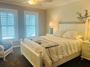 Un dormitorio con cama, dos ventanas, una mesita de noche, una lámpara y una silla.