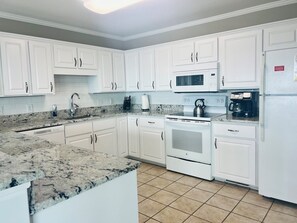 A kitchen with white cabinets, a granite countertop, a stainless steel kettle, a coffee maker, and a microwave. - Elegant Egret 4 Bedroom Home by RedAwning (Supply)