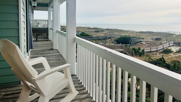 A porch with a white chair, a view of the beach, and a wooden deck. - Elegant Egret 4 Bedroom Home by RedAwning (Supply)