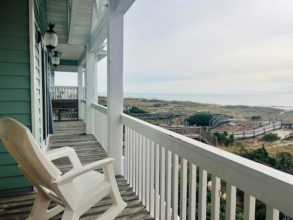 A porch with a white chair, a view of the beach, and a wooden deck.