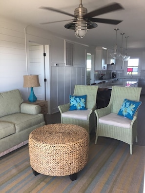 A living room with wicker furniture, a ceiling fan, and a kitchen in the background. - Almost Heaven 4 Bedroom Home by RedAwning (Supply)