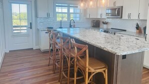 A modern kitchen with a large island, wooden chairs, white cabinets, and a pendant light fixture.