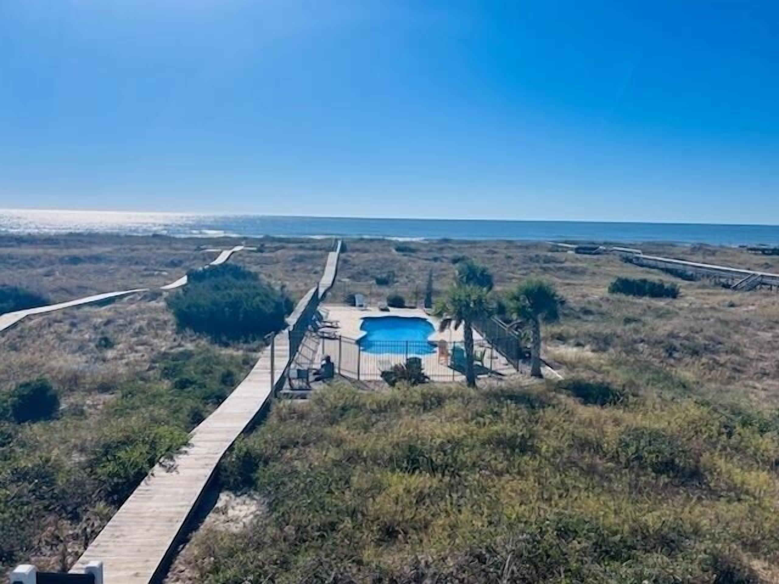A beachside property with a wooden boardwalk leading to a swimming pool surrounded by palm trees and sparse vegetation.