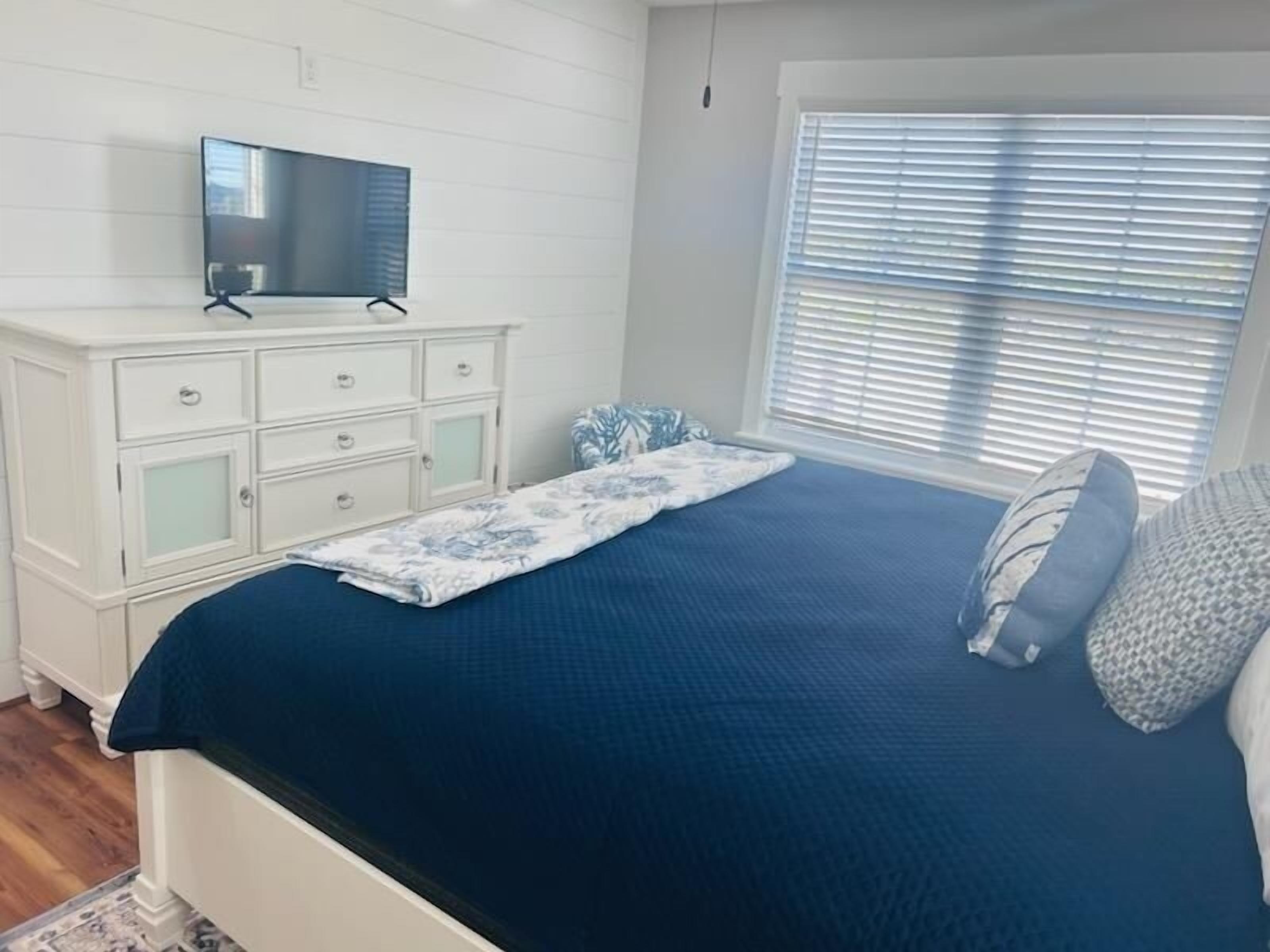 A bedroom with a bed, a dresser, a television, and a window with blinds.