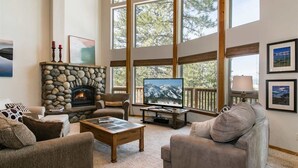A cozy living room with a stone fireplace, comfortable seating, a large TV, and large windows offering a view of trees. - Northstar Conifer Luxury 5 Bedroom Home by RedAwning (Truckee)