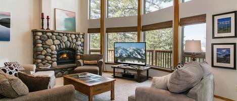 Una sala de estar acogedora con chimenea de piedra, asientos cómodos, un televisor grande y ventanas amplias con vistas a árboles.