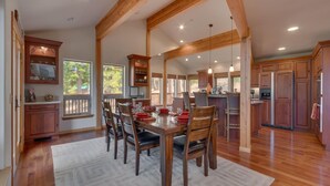 A spacious dining area with a wooden table and chairs, a kitchen with wooden cabinets and appliances, and large windows allowing natural light. - Northstar Conifer Luxury 5 Bedroom Home by RedAwning (Truckee)