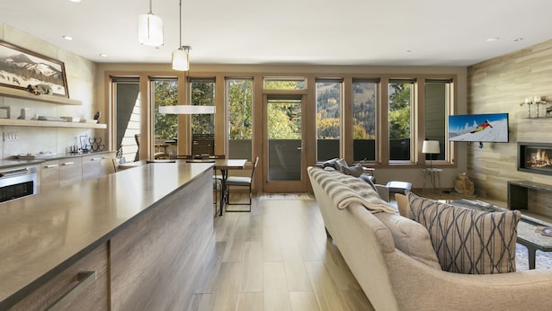 A modern kitchen with a large island, a dining area with a table and chairs, a living room with a sofa and a fireplace, and large windows with a view of trees. - Sunvail S 24d 3 Bedroom Condo by RedAwning (Vail)