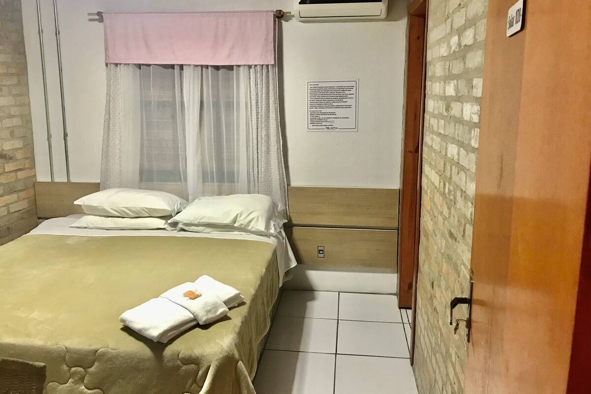 A hotel room with a bed, a window with sheer curtains, an air conditioning unit, and a door with a sign.