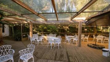 Un patio al aire libre con sillas y mesas de metal blanco, cubierto por una pérgola de madera con paneles de vidrio.