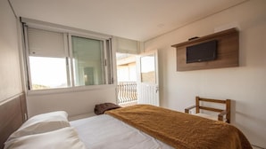 Un dormitorio con una cama, una silla, un televisor montado en la pared y una ventana con persianas.