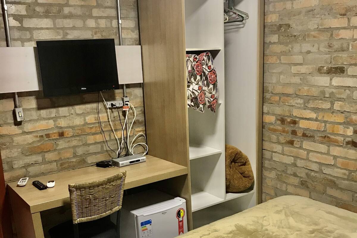 A bedroom with a bed, a desk, a television, a wardrobe, and a brick wall.