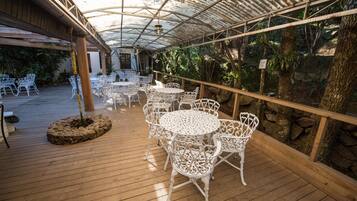 Une terrasse extérieure avec des tables et des chaises en métal blanc, entourée de rampes en bois et d’une végétation tropicale.