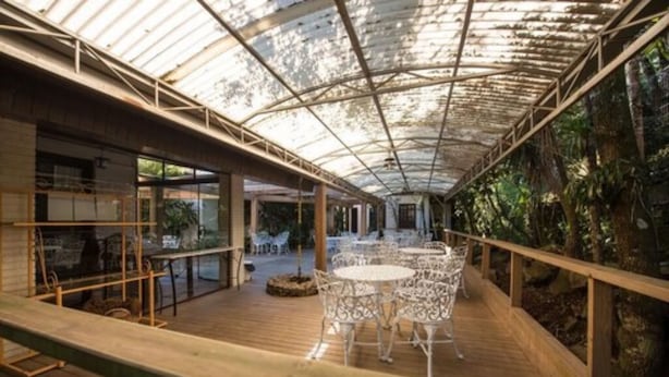 Un espacio interior con cubierta de madera, mesas y sillas de metal blanco, rodeado por paredes de vidrio y una estructura de techo metálico.
