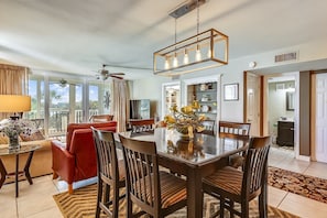 A dining area with a wooden table, chairs, and a chandelier. There is a red sofa and a TV in the background. - DP 202C Recently renovated 2 bedroom condo near Destin Harbor and amazing views by RedAwning (Destin)
