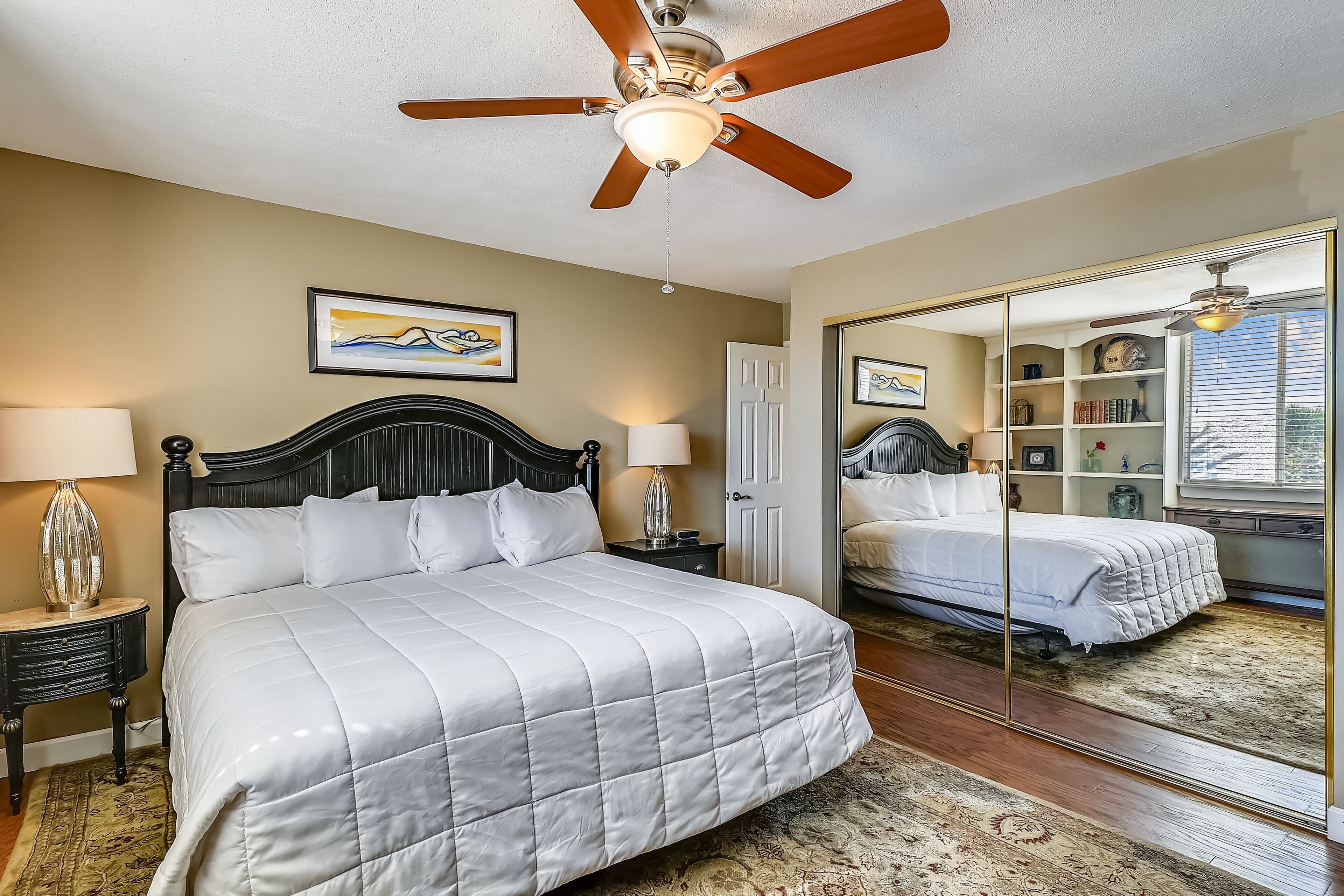 A bedroom with a bed, a ceiling fan, a nightstand with a lamp, a framed picture on the wall, and a large mirror reflecting the room.