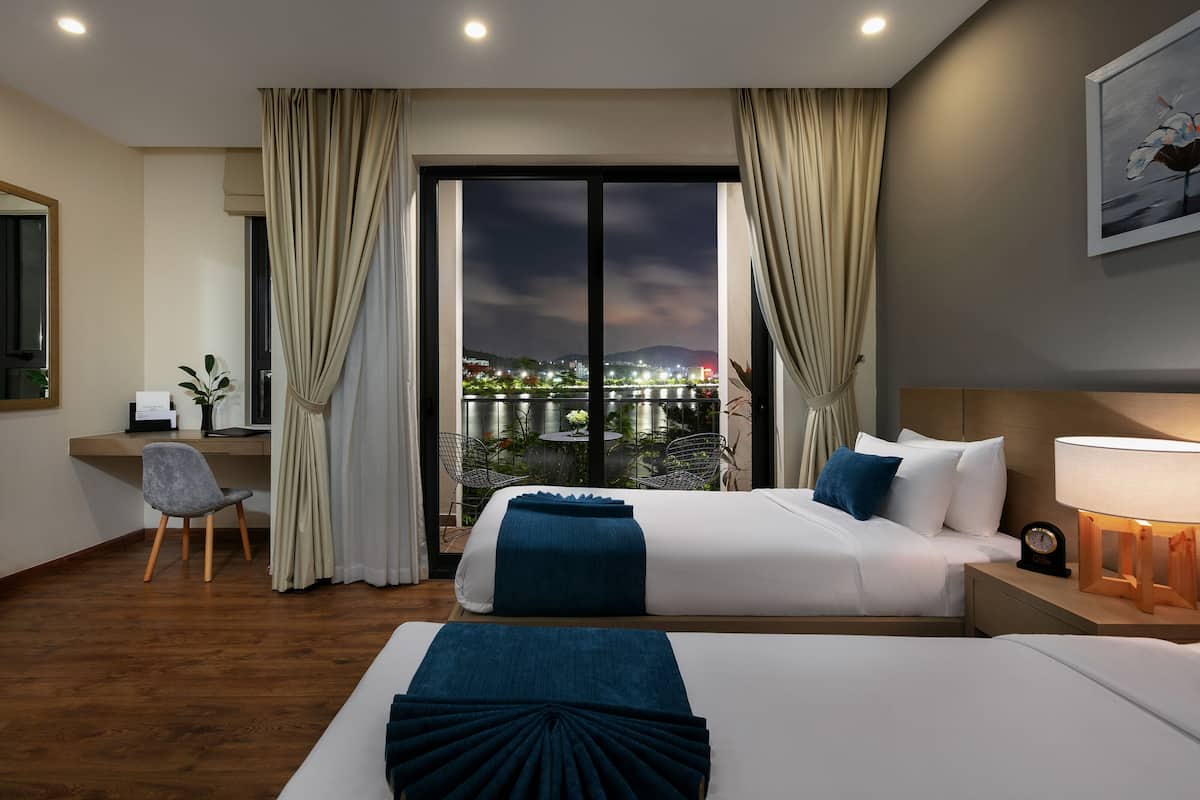 A hotel room with a bed, a desk, a chair, and a view of a cityscape through a large window.