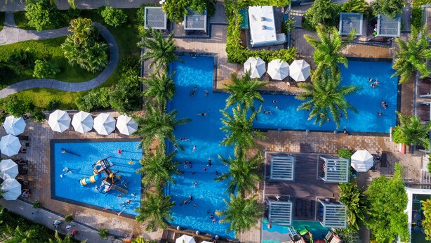 An aerial view of a resort with multiple swimming pools surrounded by palm trees and sun loungers. - La Batisse Resort Halong (Bai Chay)