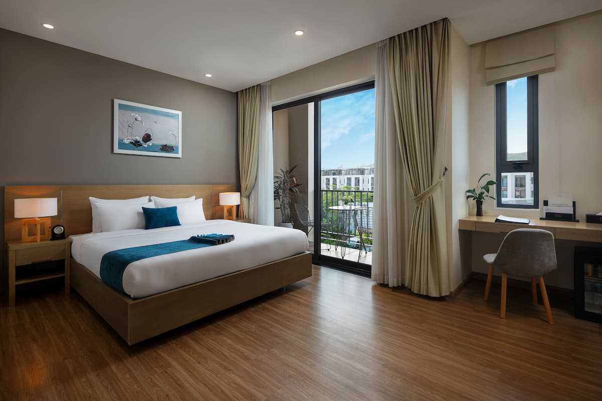 A modern hotel room with a large bed, a desk with a computer, and a balcony with a view of the city.