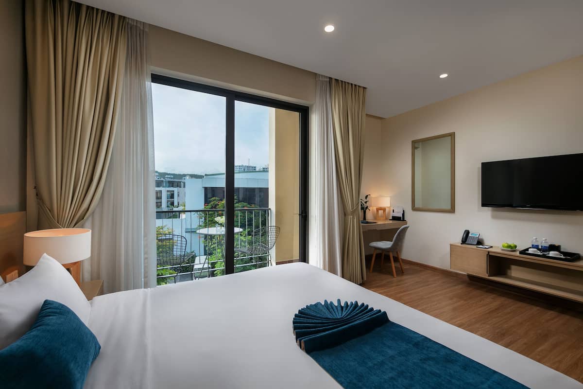 A hotel room with a large bed, a desk, a TV, and a balcony with a view of buildings.