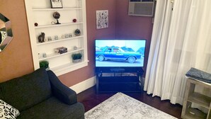 Een woonkamer met een bank, een televisie waarop een auto wordt getoond, een plank met decoratieve objecten, en een raam met gordijnen.