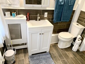 Une salle de bain avec un lavabo blanc, des toilettes et un meuble de rangement. Une serviette bleue est accrochée au mur, ainsi qu’une poubelle et un rideau de douche.