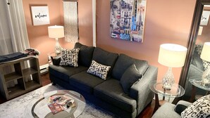 Een woonkamer met een donkergrijze bank, gestreepte kussens, een glazen salontafel met tijdschriften, een wandlamp en een groot abstract schilderij.