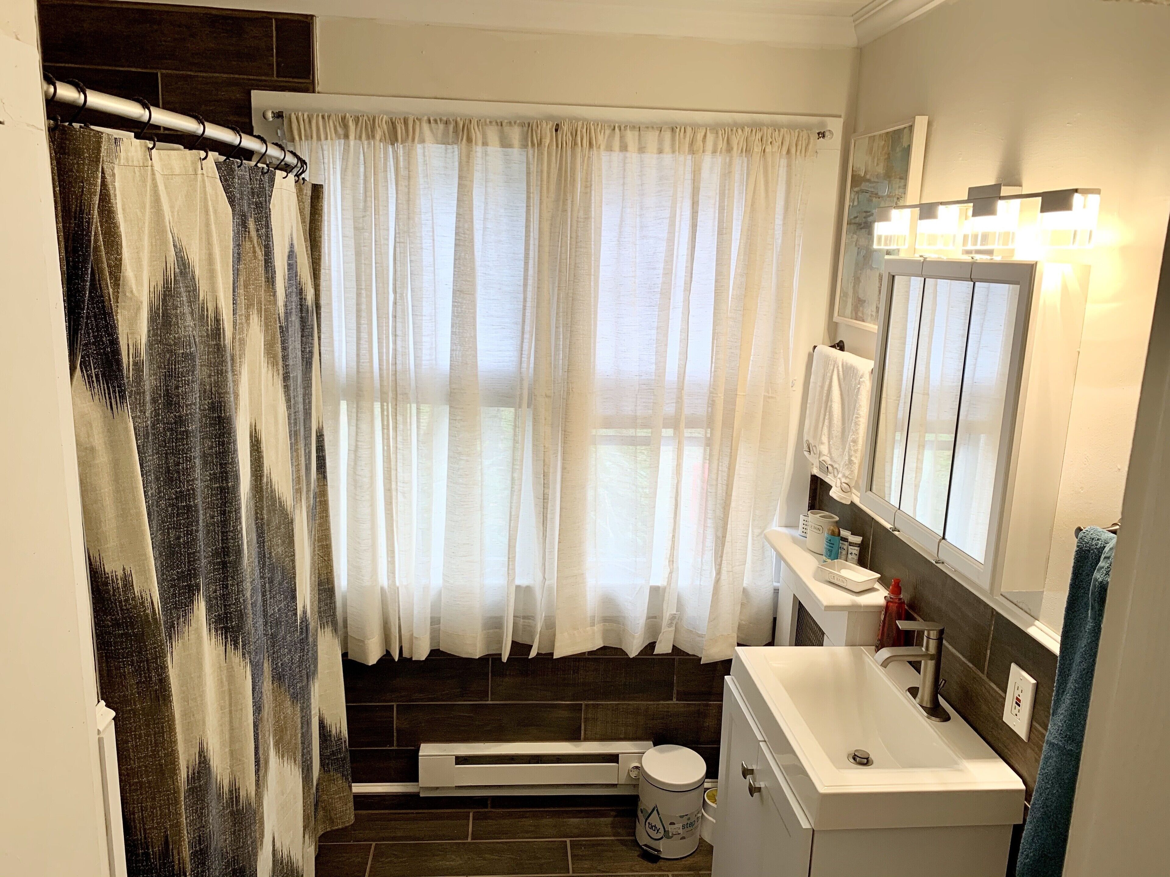 A bathroom with a shower curtain, a sink, a mirror, and a window with curtains.