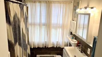 A bathroom with a shower curtain, a sink, a mirror, and a window with curtains.